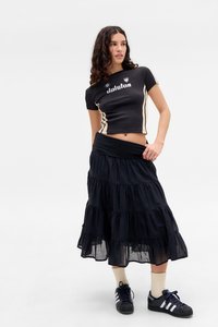 Noor noor, kellel on lainelised juuksed, kannab musta lühikeste varrukatega t-särki, kihilist musta seelikut, kreemi värvi sokke ja musti Adidas tossusid, poseerib valge tausta ees.