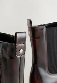 Skórzane botki za kostkę w ciemnym brązie o gładkim wykończeniu, z elastycznymi panelami bocznymi, detalami szwów oraz metalowymi akcentami na obcasie.