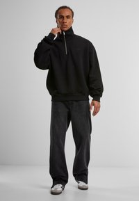 Svart zip-up hettegenser med høy krage, avslappet passform og ribbestrikkede mansjetter; paret med mørke vide bukser og hvite joggesko.