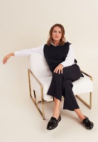 Gilet sans manches bleu marine sur une chemise blanche, associé à un pantalon large noir et des mocassins noirs avec des accents cloutés, assis sur une chaise blanche.