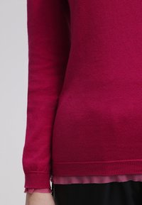 Gros plan d'une personne portant un pull ajusté à manches longues magenta sur une chemise rose plus claire, avec un pantalon noir visible en bas.