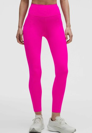 Close-up van een persoon die felroze leggings met hoge taille en witte sportschoenen draagt tegen een effen achtergrond.