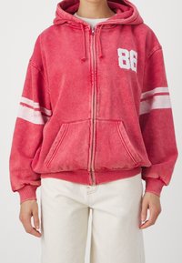 Sudadera con cremallera roja, con rayas blancas y el número "86" en el frente. Fabricada con material suave, cuenta con bolsillos frontales y puños acanalados.