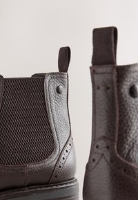 Brune læderankelstøvler med mesh-paneler, kontrastsyninger og små trækflapper; har runde tæer og mønstrede teksturer.