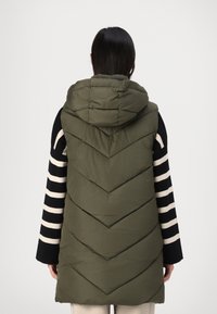 Olivgrön pufferväst med en chevron-design, med huva och svartvita randiga ärmar. Slät textur, medellängd.
