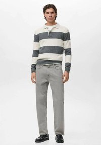 Hombre vestido con un suéter polo de rayas blancas y grises de manga larga, jeans rectos de color gris claro y zapatos de cuero negros, de pie contra un fondo blanco.