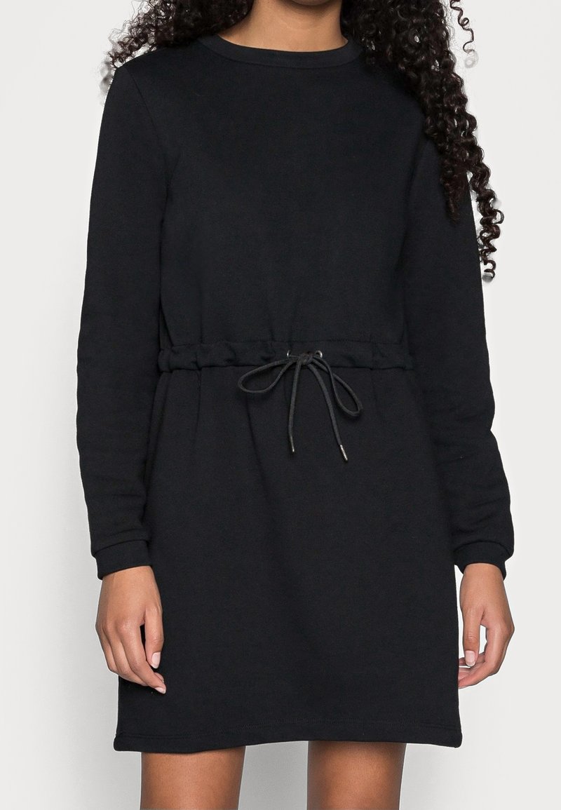 Personne portant une robe noire à manches longues avec un col rond et une taille à cordon noué devant.