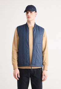 Colete acolchoado azul-marinho com fecho éclair e um bolso no peito, usado sobre uma camisa de manga longa beige. Jeans escuros e um boné azul-marinho completam o visual.