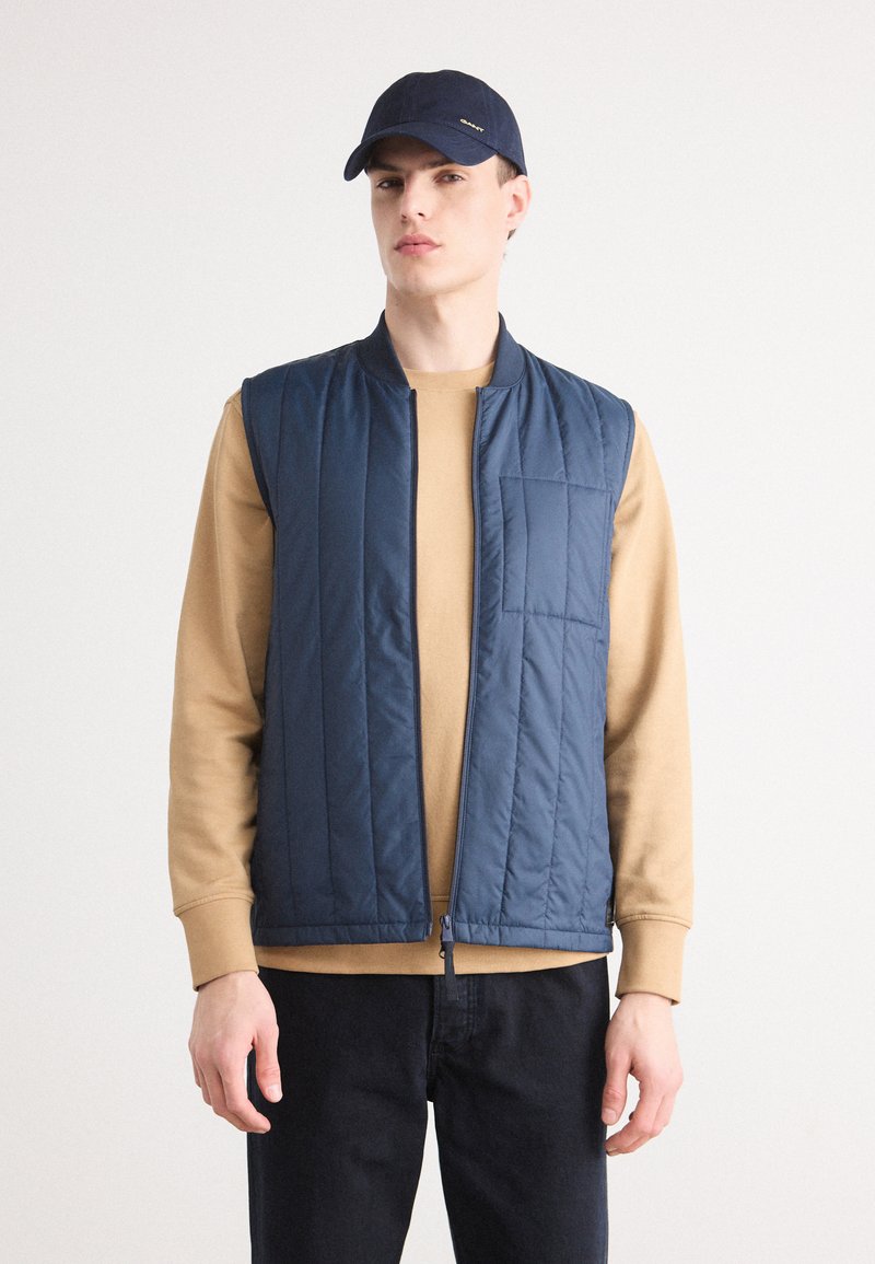 Colete acolchoado azul-marinho com fecho éclair e um bolso no peito, usado sobre uma camisa de manga longa beige. Jeans escuros e um boné azul-marinho completam o visual.