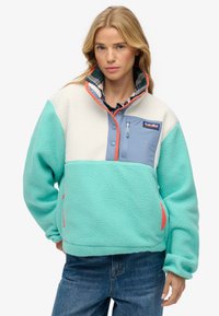 Femme portant un pull polaire bleu sarcelle et crème avec une poche poitrine bleue et des détails de fermeture éclair orange, associée à un jean bleu.