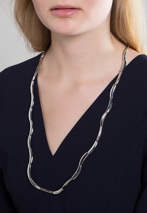 Collana in metallo argento con anelli allungati e ondulati. Il design è liscio con una finitura lucida, indossata sopra un capo con scollatura a V profonda.