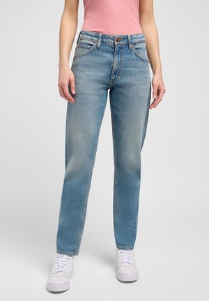 Kvinde iført lyseblå jeans med lige ben, hvide sneakers og en lyserød ærmeløs top, stående mod en ensfarvet baggrund.