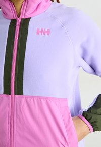 Lillakas fliisjakks roosa ja rohelise aktsentidega, millel on ees sü zip, kõrge kaelus ja reguleeritavad mansetid. Logo rinnal.