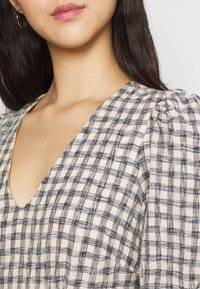V-ringad blus i krämfärg med svart rutmönster. Har puffärmar och strukturerad tyg, vilket skapar ett mjukt, lager-på-lager utseende.