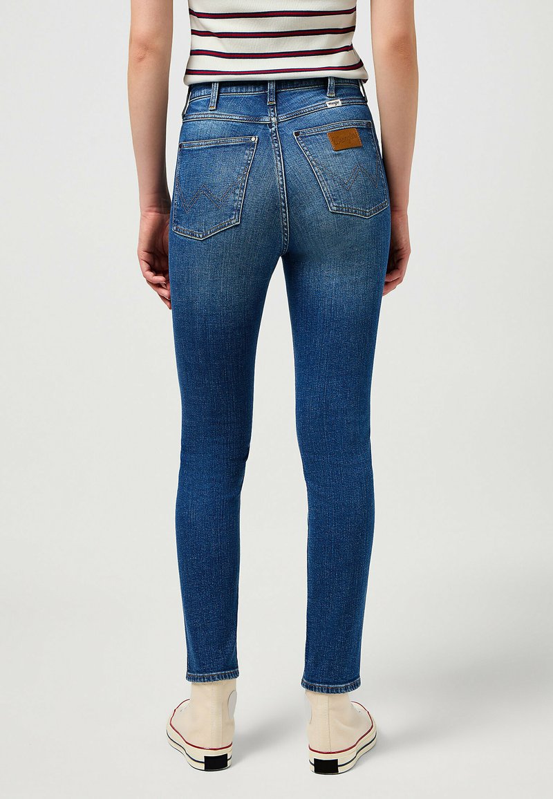 Skinny jeans made of blue denim, featuring a high-rise fit, back pockets, and a leather logo patch on the waistband. Textured surface.