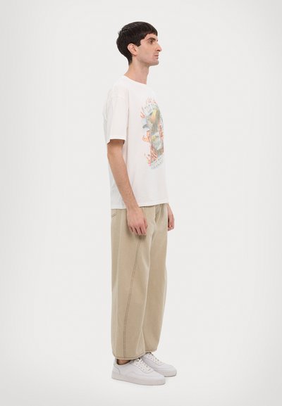 T-shirt graphique blanc avec un design coloré, associé à un pantalon large beige et des baskets blanches, montré de côté.
