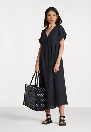 Femme portant une robe noire à col en V tenant un grand sac à motifs, avec des sandales noires et un bracelet en or ainsi que des boucles d'oreilles.