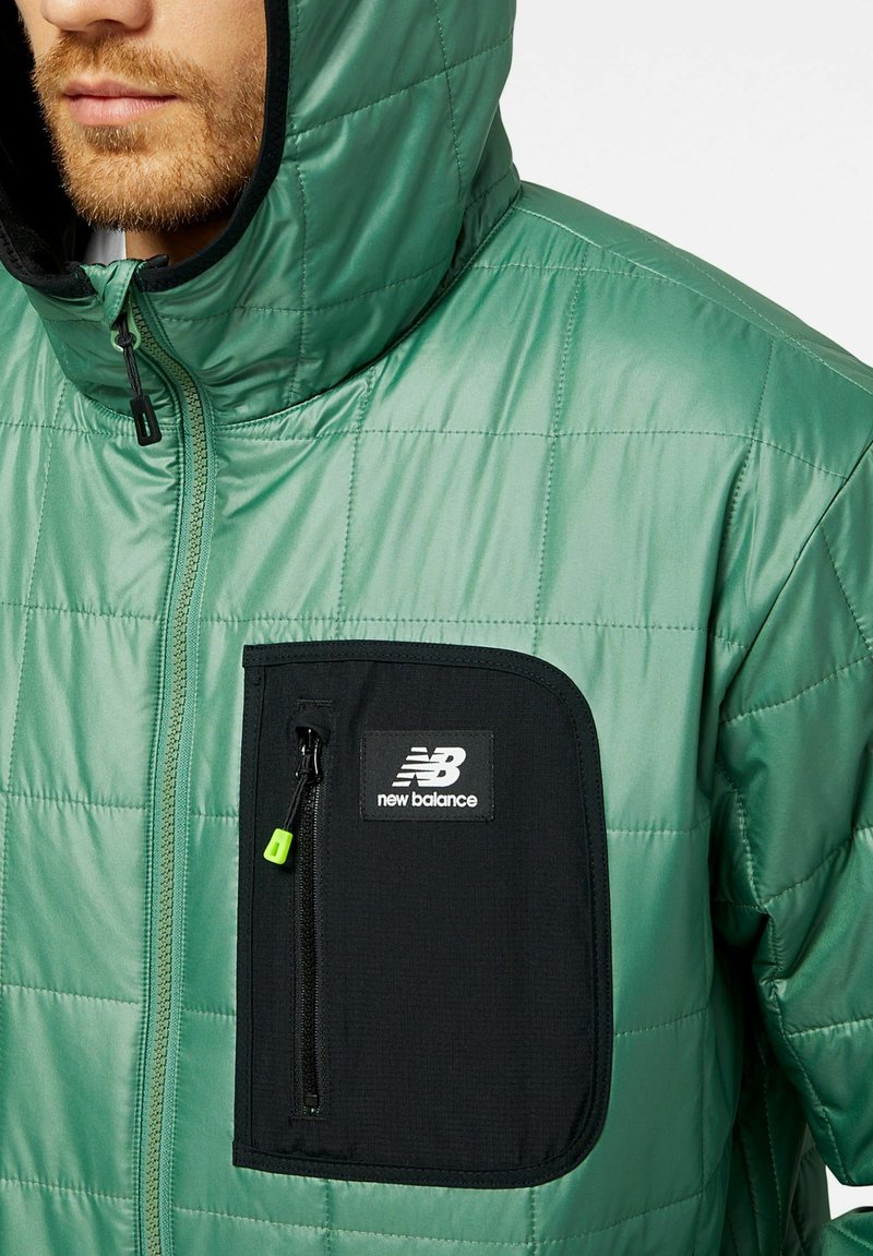 Grüne gesteppte Jacke mit einer schwarzen Reißverschlusstasche, die ein Logo trägt. Hergestellt aus glattem, glänzendem Material mit einer Kapuze und kontrastierenden Akzenten.
