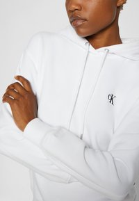 White hoodie with a drawstring, featuring an embroidered logo in black. Soft texture, ribbed cuffs, and a flexible fit.