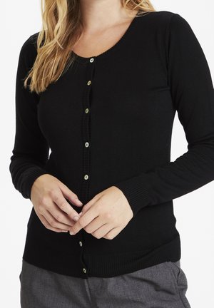 Femme portant un cardigan noir à manches longues boutonné et un pantalon gris, les mains doucement jointes à la taille.