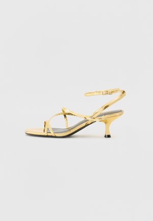 Zlate sandale s trakami in tanjšim petam, kovinskim finišem in trakovi okrog gležnja. Imajo križno zasnovo z osnovnim črnim podplatom.