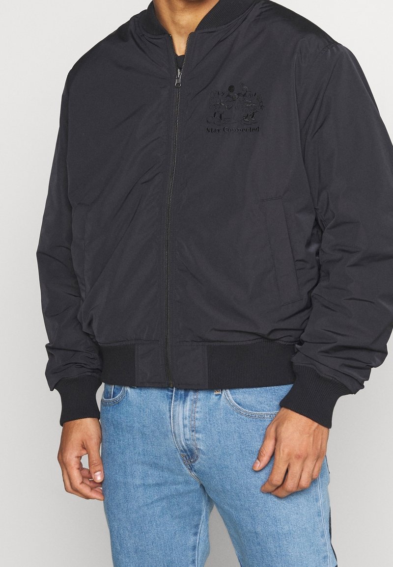 Svart bomberjacka med dragkedja, ribbade ärmslut och nederkant, med ett präglat "Stay Connected"-logotyp på bröstet. Stylerad över blå jeans.