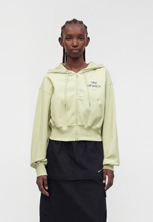 Mladá žena s copánky, která nosí světle zelenou mikinu Nike Athletics na zip a černou sukni s malým bílým logem Nike.