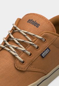 Baskets en toile beige avec lacets à motifs blancs et noirs, marquées "etnies" sur la languette et le côté, semelle beige avec une bande noire.