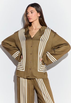 Vrouw staat met handen in de zij, draagt een olijfgroen knoopshirt en broek met witte geometrische streeppatronen.