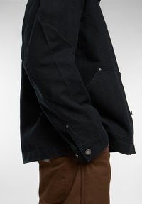 Zwarte canvasjas met een licht gestructureerd ontwerp, knoopaccenten en klepzakken; gecombineerd met een bruine broek, zijaanzicht.