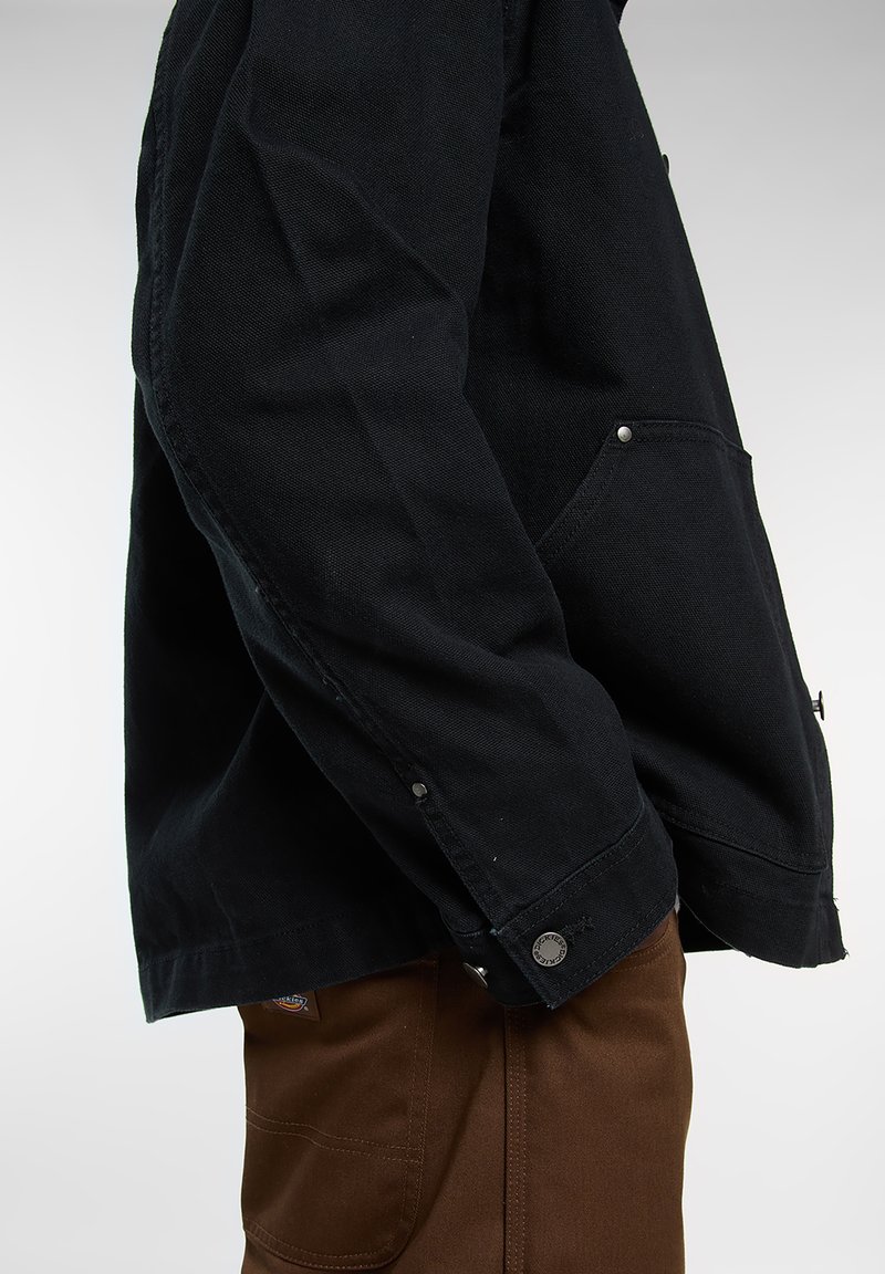 Giacca nera in tela con design leggermente strutturato, dettagli con bottoni e tasche con pattina; abbinata a pantaloni marroni, vista laterale.