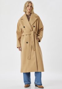 Beige dobbeltrækket frakke med bælte, brede lapelkraver og store knapper. Har sidelommer og en oversized pasform.