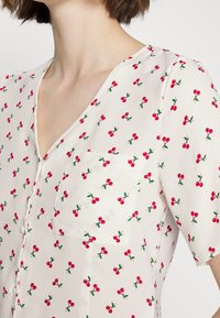 Blusa de mangas curtas em tecido creme, com um padrão de cerejas em vermelho e verde. Detalhes notáveis incluem um decote em V e um bolso no peito.