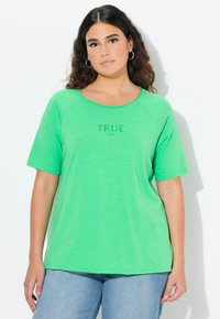 Donna con capelli ricci scuri che indossa una maglietta verde a maniche corte con la scritta "TRUE" e jeans blu chiaro, in piedi contro uno sfondo bianco.