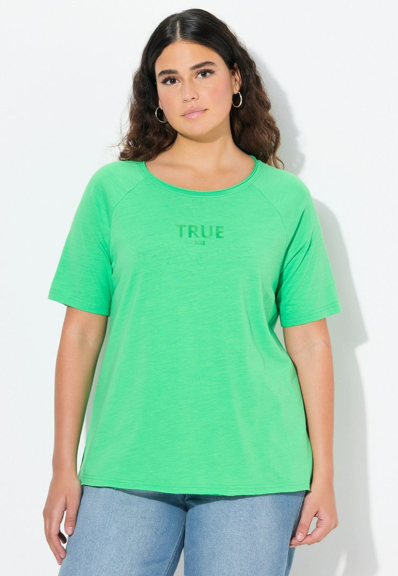 Donna con capelli ricci scuri che indossa una maglietta verde a maniche corte con la scritta "TRUE" e jeans blu chiaro, in piedi contro uno sfondo bianco.