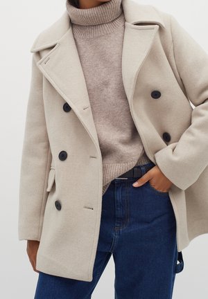 Person iført beige dobbeltradet frakke over lysebrun rullekrave og mørkeblå jeans med den ene hånd i lommen.