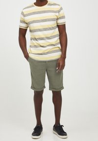 T-shirt a maniche corte a righe gialla, grigia e bianca con colletto rotondo; abbinata a pantaloni corti khaki arrotolati e sneakers nere.