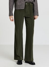 Pantalon large en velours côtelé vert avec nervures verticales, taille haute et fermeture à boutons, assorti à un cardigan gris et des chaussures noires.