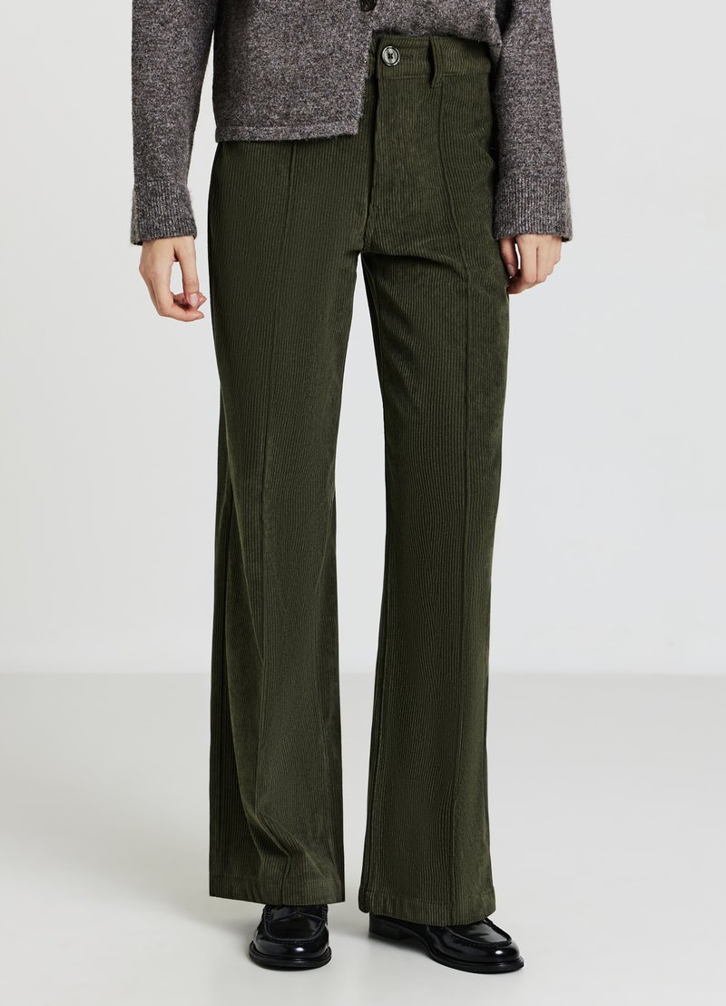 Pantalon large en velours côtelé vert avec nervures verticales, taille haute et fermeture à boutons, assorti à un cardigan gris et des chaussures noires.
