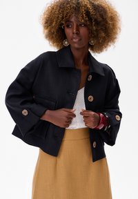 Czarna, teksturowana kurtka o pudełkowym fasonie, z dużymi drewnianymi guzikami i rękawami do trzech czwartej. W zestawieniu z białym topem i musztardową spódnicą.