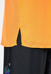 Orange Chiffon-Bluse mit seitlichen Schlitzen, kombiniert mit schwarzen Hosen, die am Saum ein farbenfrohes Blumenmuster haben. Weicher Stoff, leichtes Material.