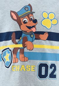 T-shirt gris avec un chien de cartoon en uniforme de police bleu, avec des accents jaunes, des rayures noires, et un texte indiquant "CHASE 02" et une empreinte de patte.