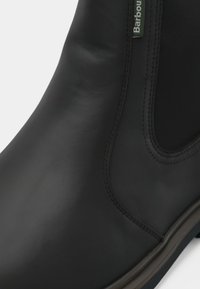 Detalhe de uma bota Chelsea de couro preto liso com painel lateral elástico e etiqueta da marca Barbour.