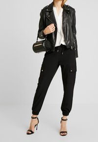 Donna con giacca di pelle nera, camicia bianca, pantaloni cargo jogger neri, sandali con tacco alto neri, che tiene in mano una piccola borsa cilindrica nera.