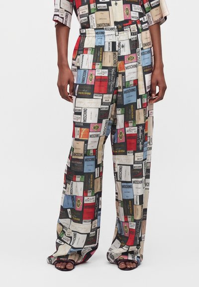 Bukser og matchende skjorte med farverigt patchworkprint med forskellige Moschino-mærkeetiketter, båret med sorte åbne sandaler.