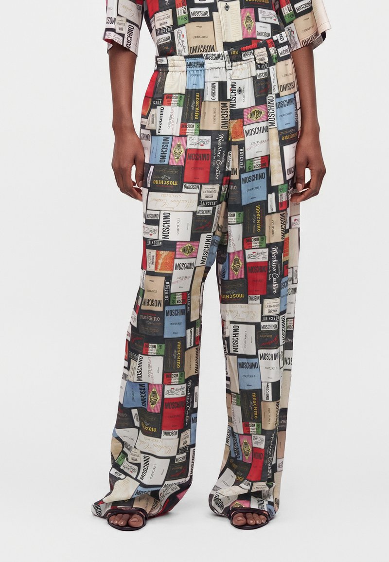 Pantalon et chemise assortie avec un imprimé patchwork coloré présentant diverses étiquettes de la marque Moschino, portés avec des sandales noires à bouts ouverts.