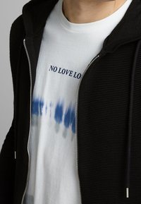 Černá mikina na zip s texturovanou tkaninou, na niž je vrstvené bílé tričko s modrošedým batikovaným vzorem a nápisem "NO LOVE LOST."