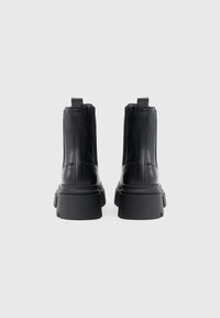 Bottines en cuir noir avec une semelle épaisse noire, présentant une texture lisse, des languettes à l'arrière et des détails de couture verticale.