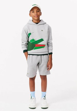 Un sweat à capuche gris avec un design graphique vert et rouge, associé à un short gris, des baskets blanches et une casquette blanche ; les deux portent des logos Lacoste.