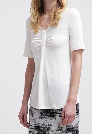 Vrouw met een wit kortermig shirt met geplooide voorkant en een zwart-witte rok met abstract patroon.